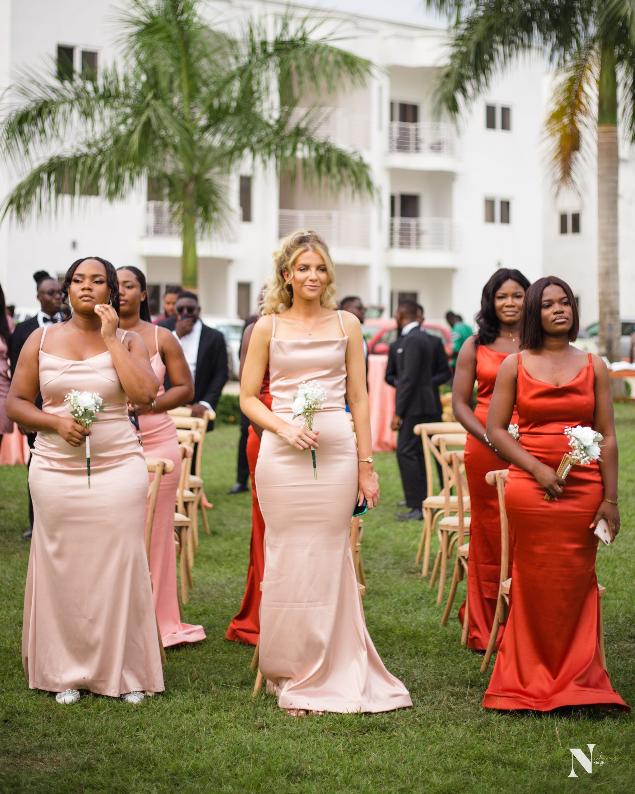 Bridesmaids in pink and red dresses walking in formation at an outdoor wedding ceremony.