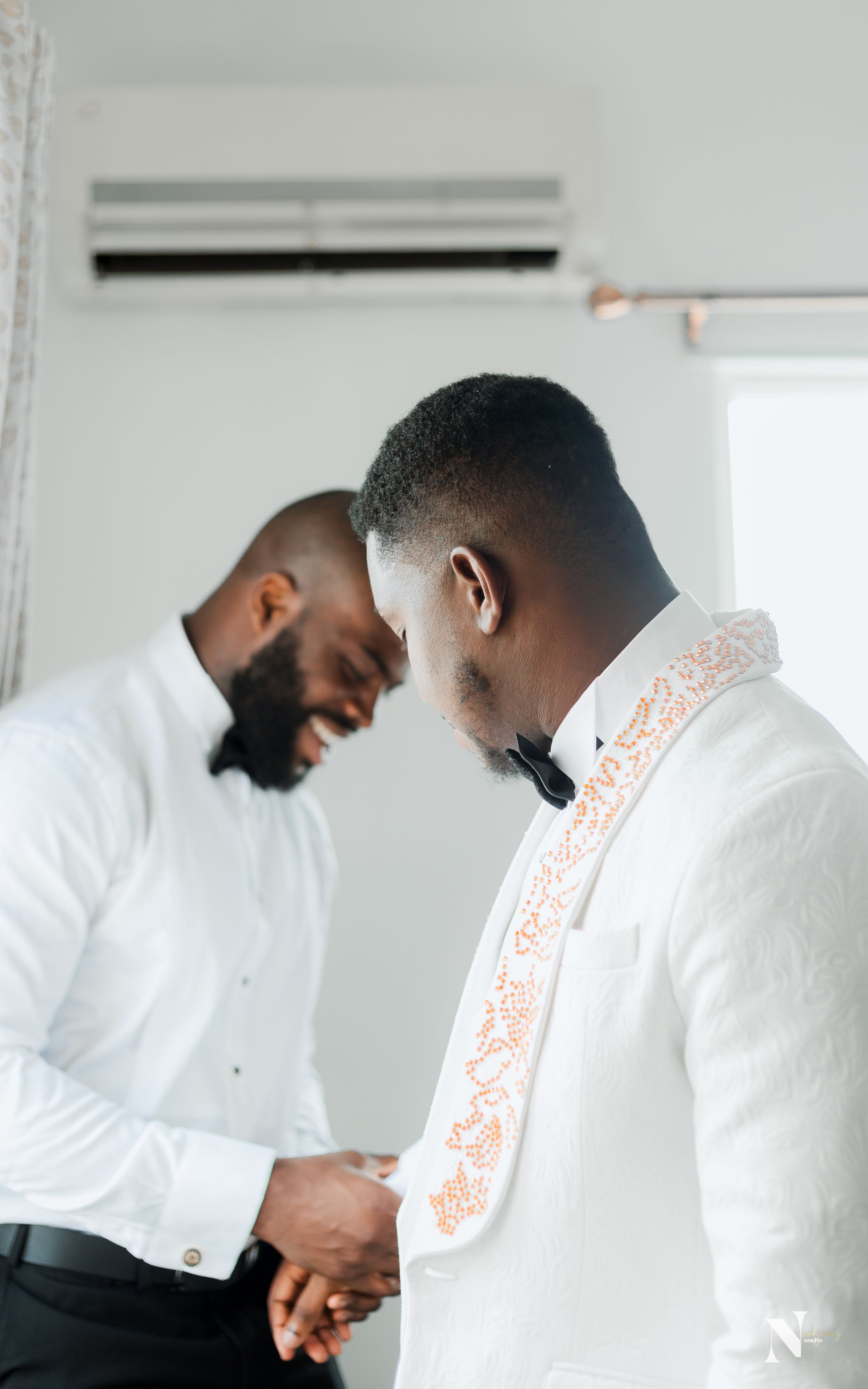 Two men in white formal wear share a candid moment during wedding preparations.