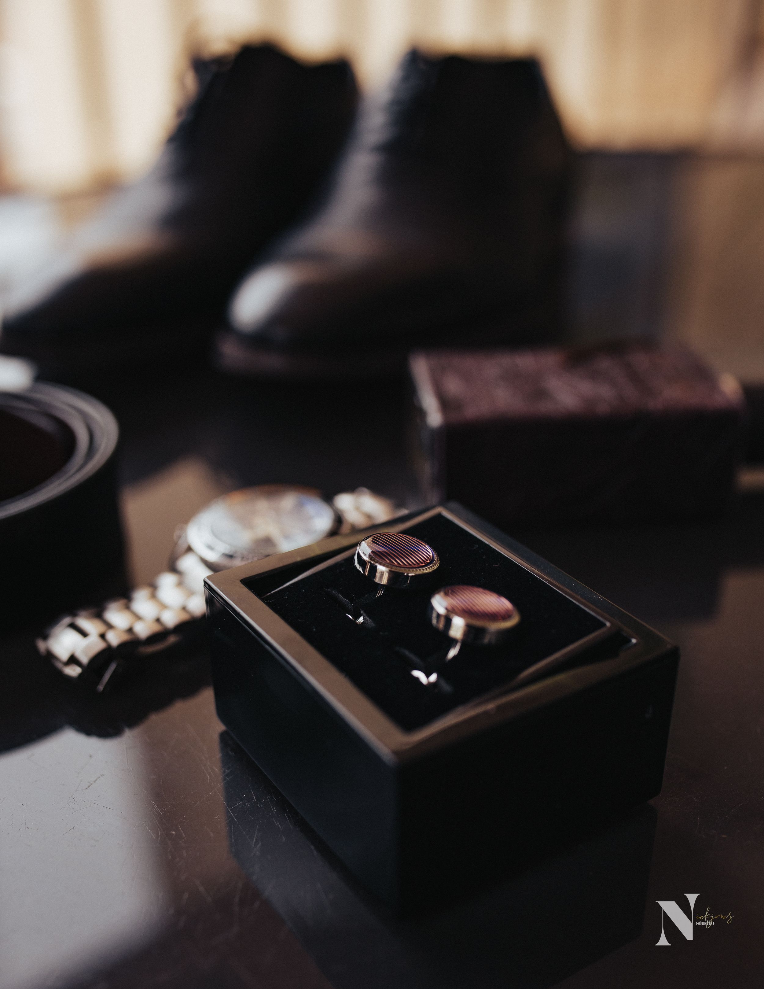 Wedding accessories including rings, watch, and dress shoes arranged on dark surface.