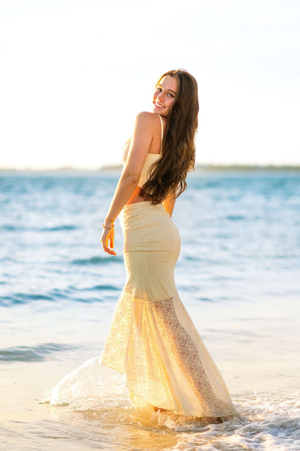 A person in a flowing cream dress twirls on the beach with ocean waves in the background.