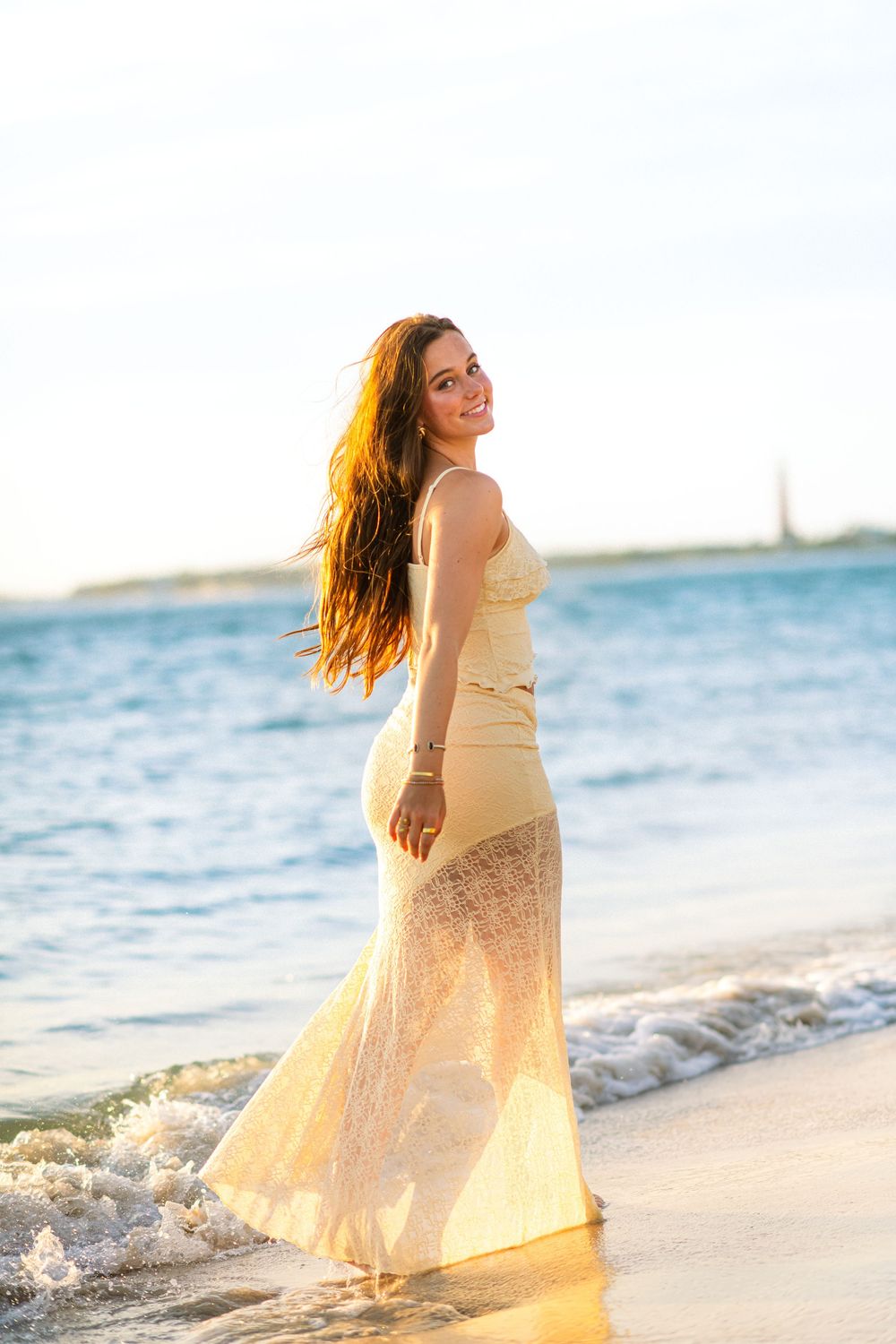 Woman in flowing cream dress dancing on beach with waves in background.