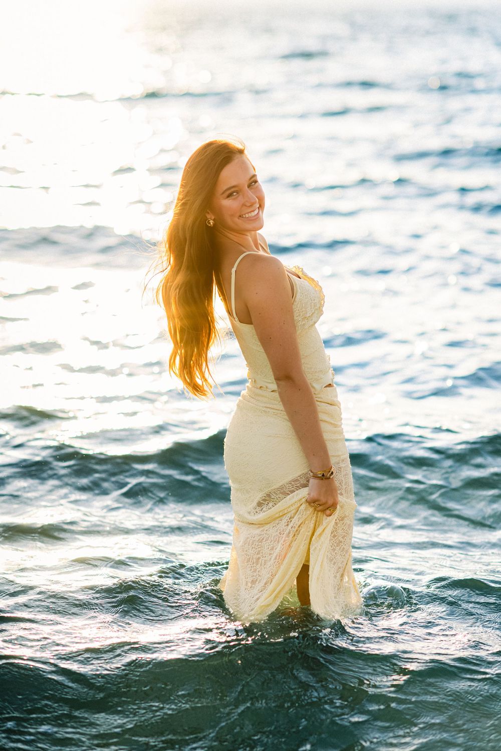 A person in a light-colored dress stands in ocean water with sunlit waves behind them.
