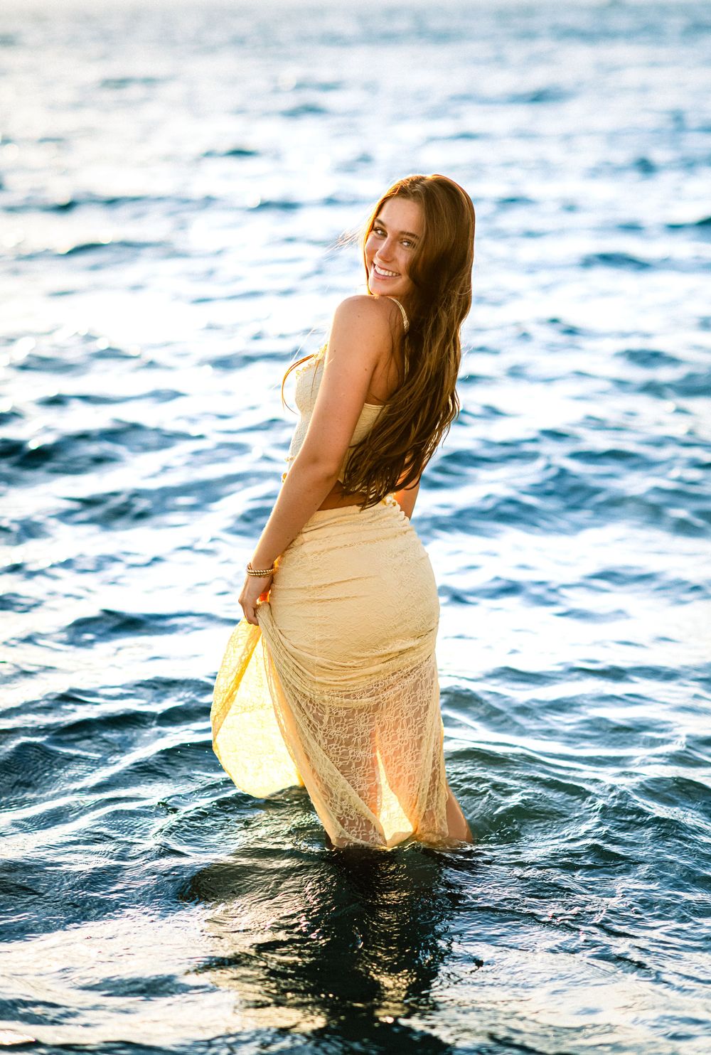 A person in a cream lace dress sits in shallow ocean waters during golden hour.