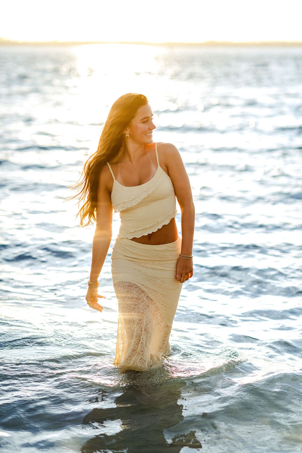 Woman in light colored dress wading through ocean water during sunset.