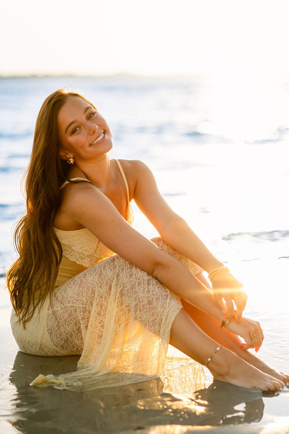 A person in lace dress sits at waters edge during sunset with waves in background.