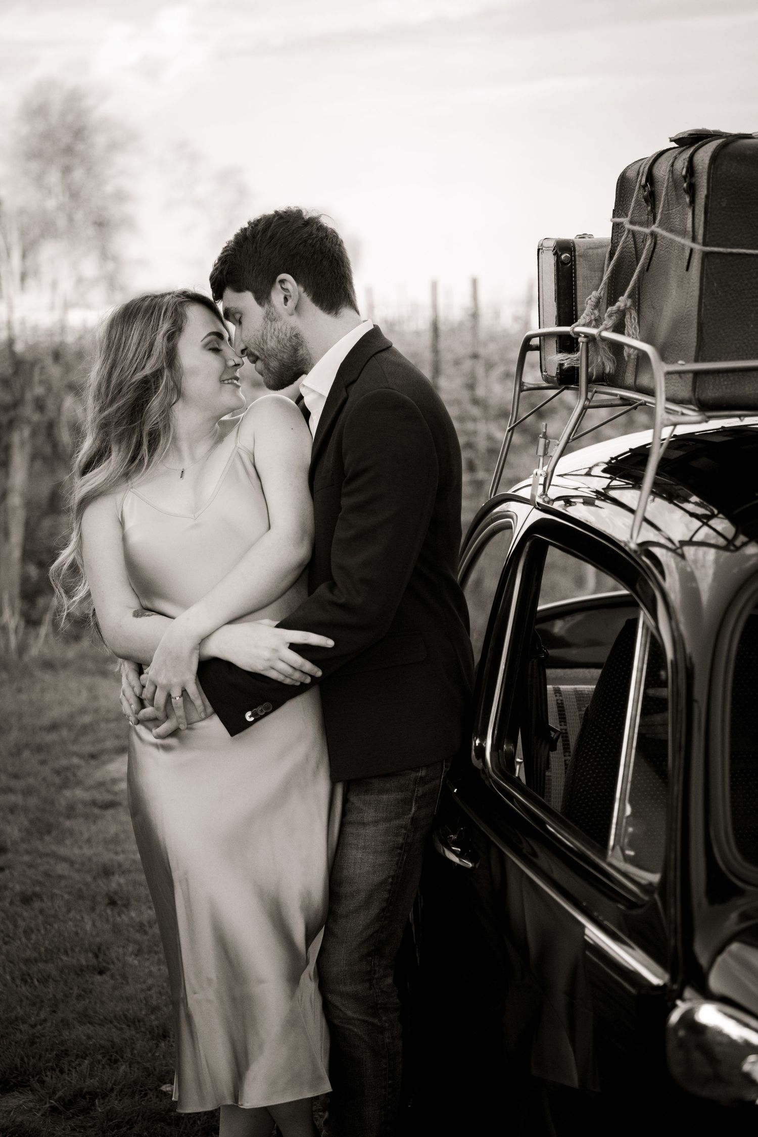 A romantic black and white moment between a couple next to a vintage car with luggage.