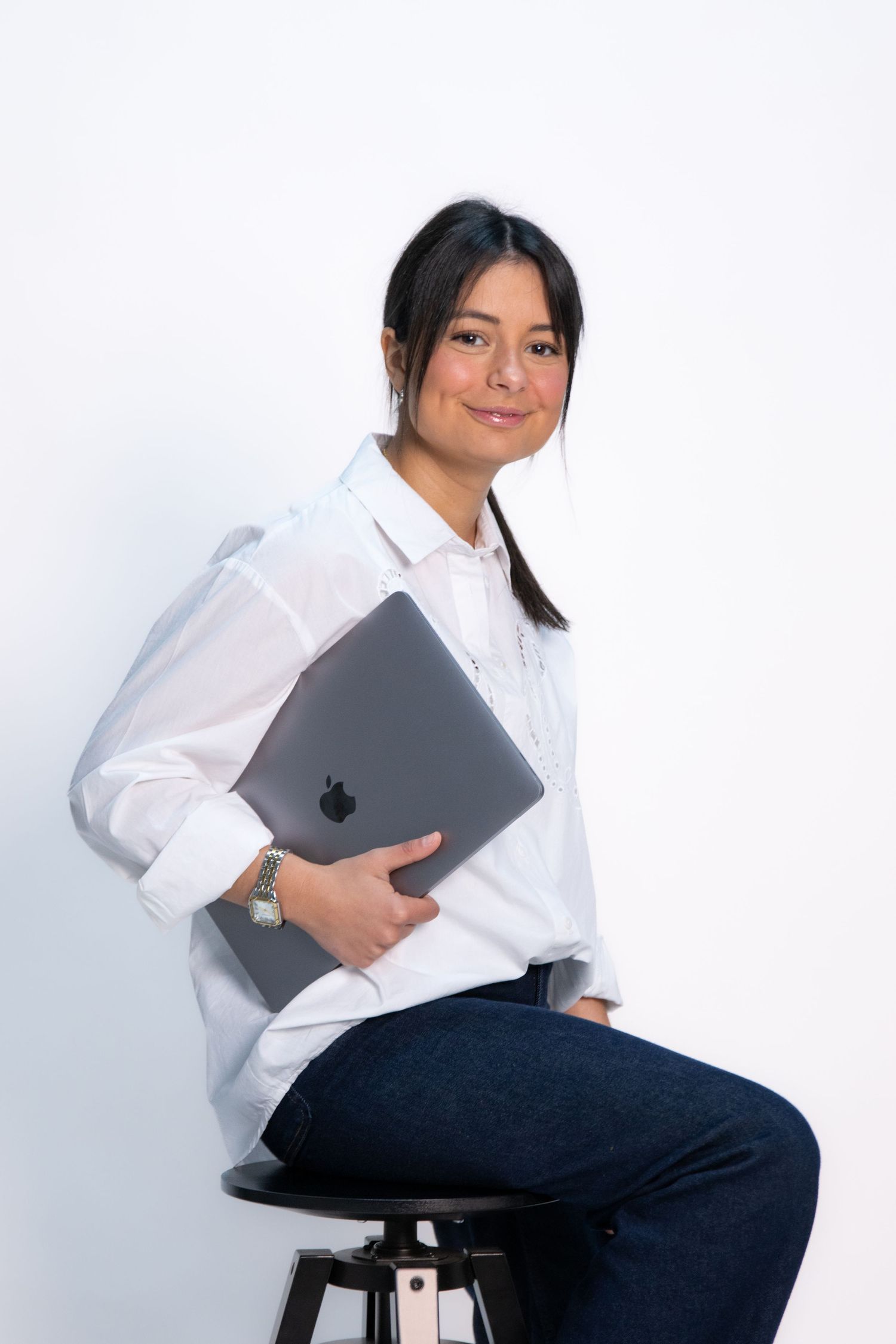 Professional wearing a crisp white button-down shirt holds a gray laptop while seated on a dark stool.