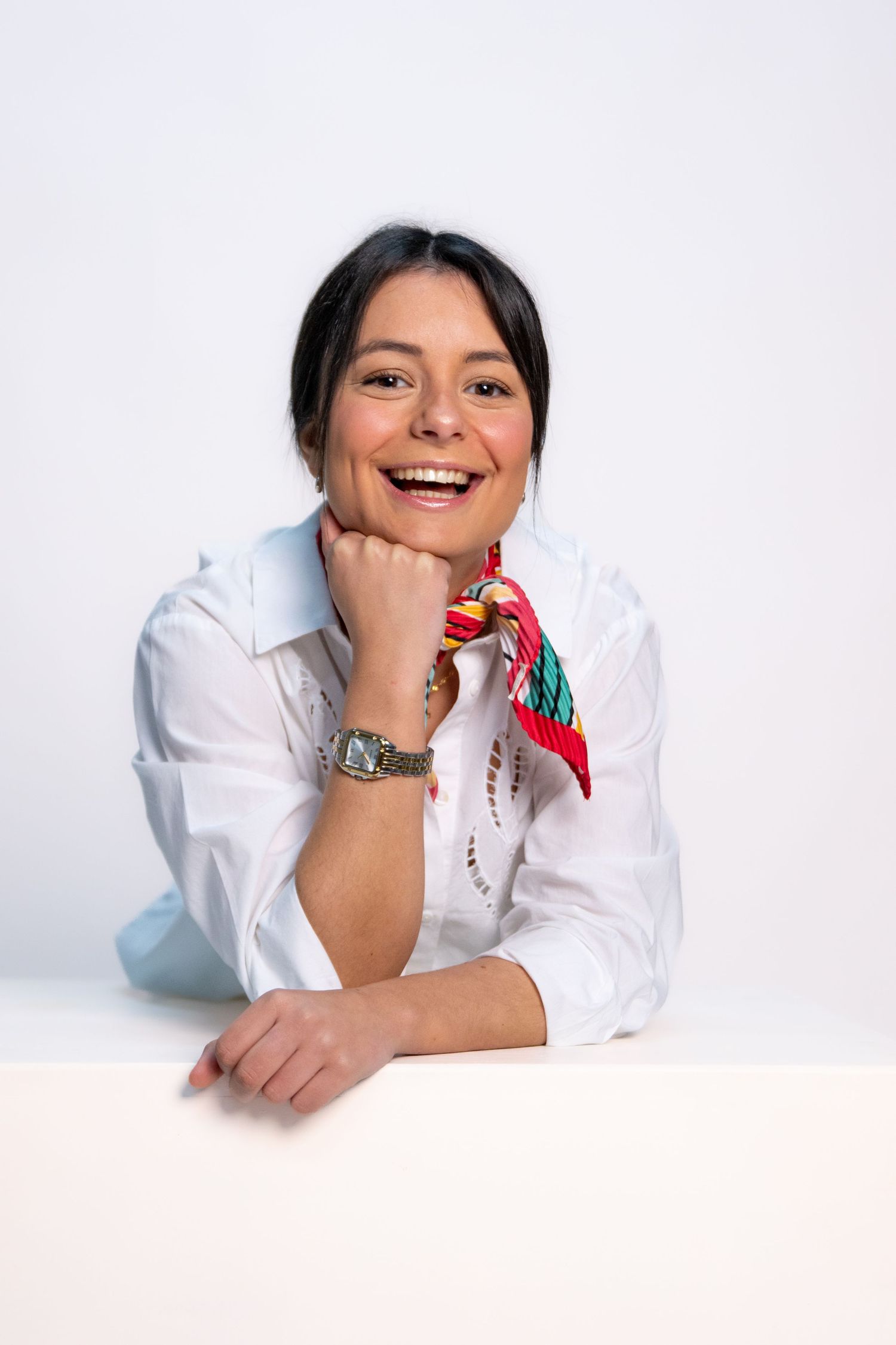 Cheerful professional in white shirt and colorful scarf rests chin on hand while posing against white background.