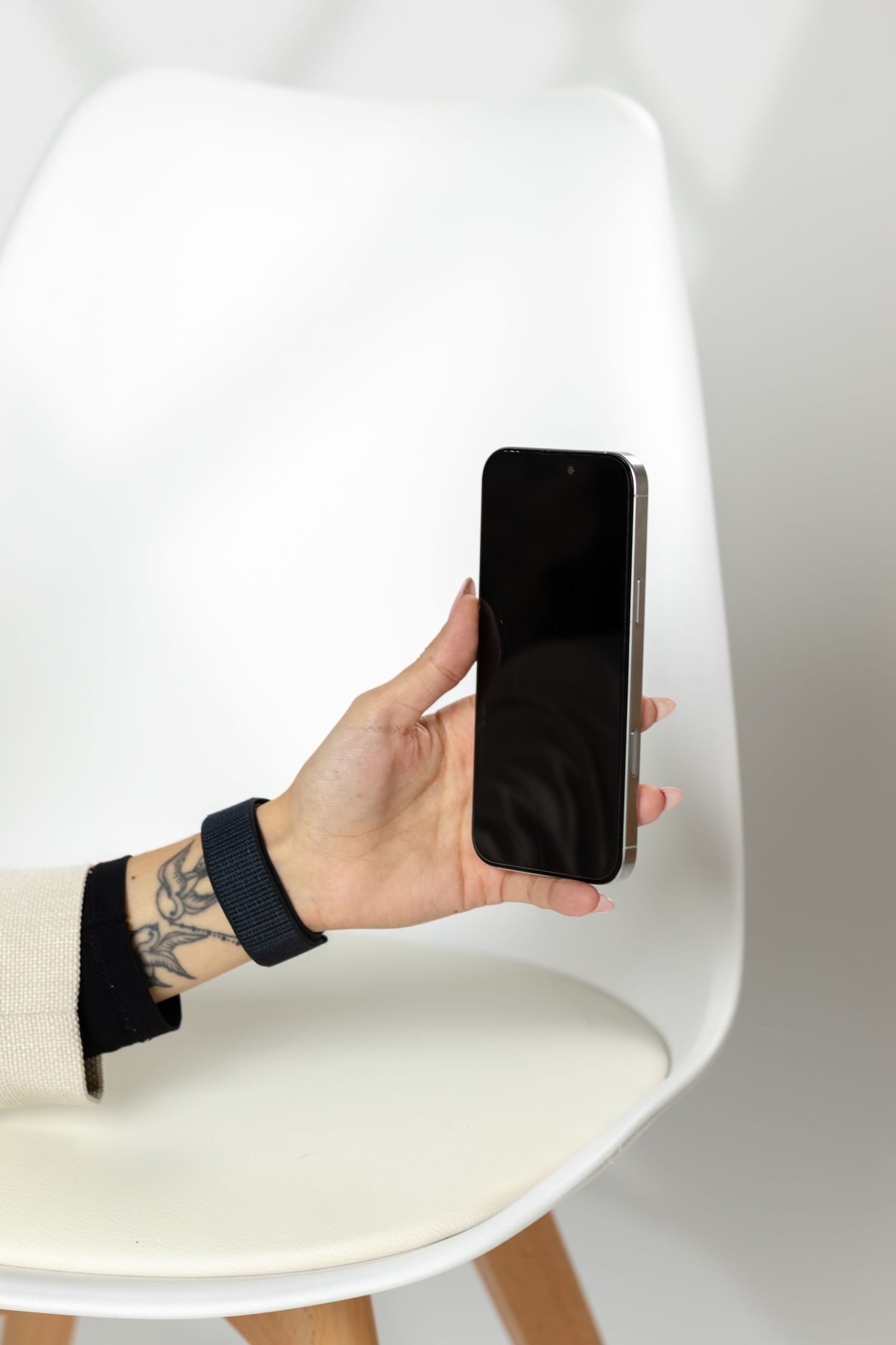 A tattooed hand holding a black smartphone against a white background with a modern chair visible.