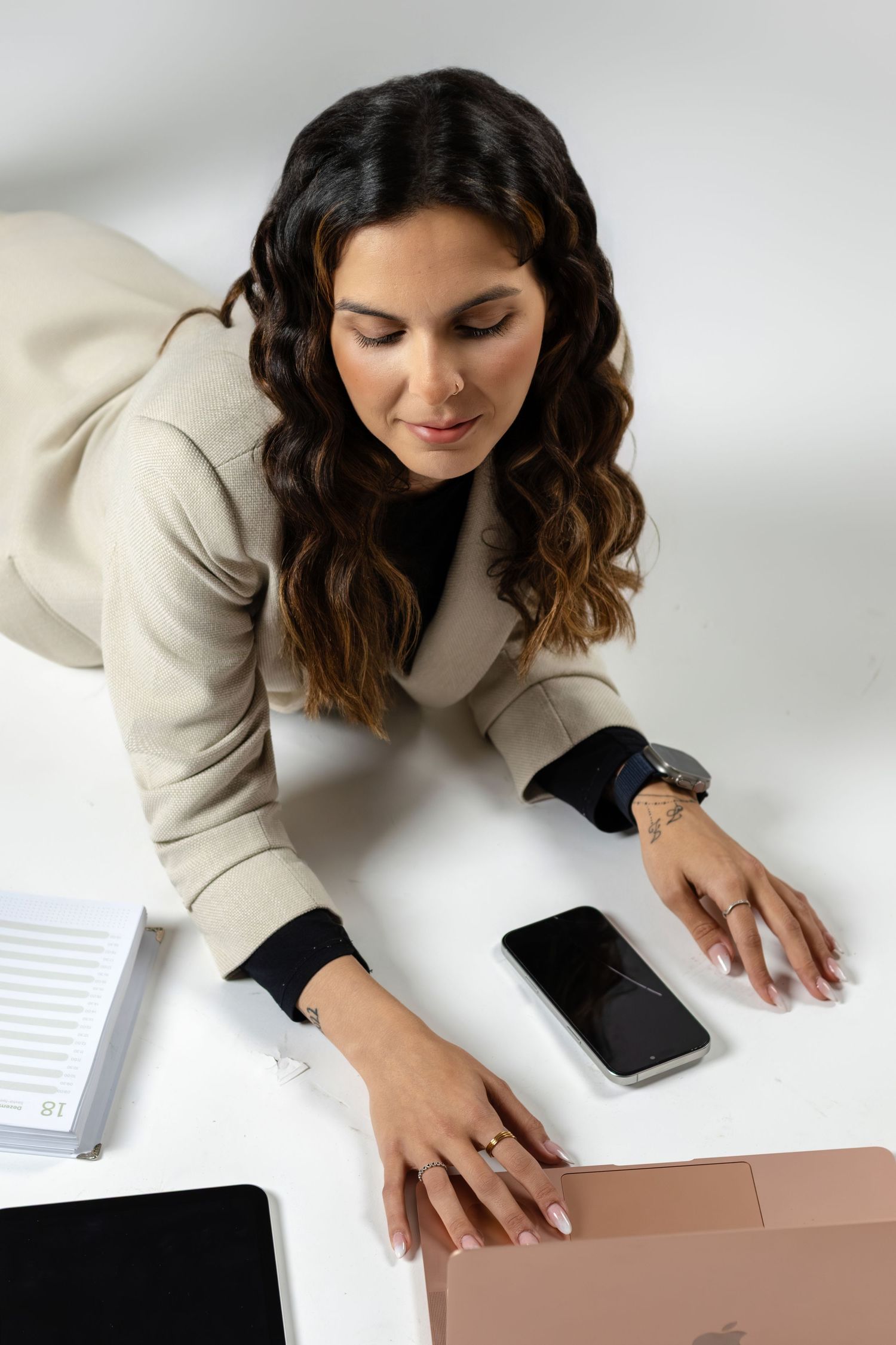 Professional working at white desk with laptop, smartphone and documents, wearing beige blazer and black top.