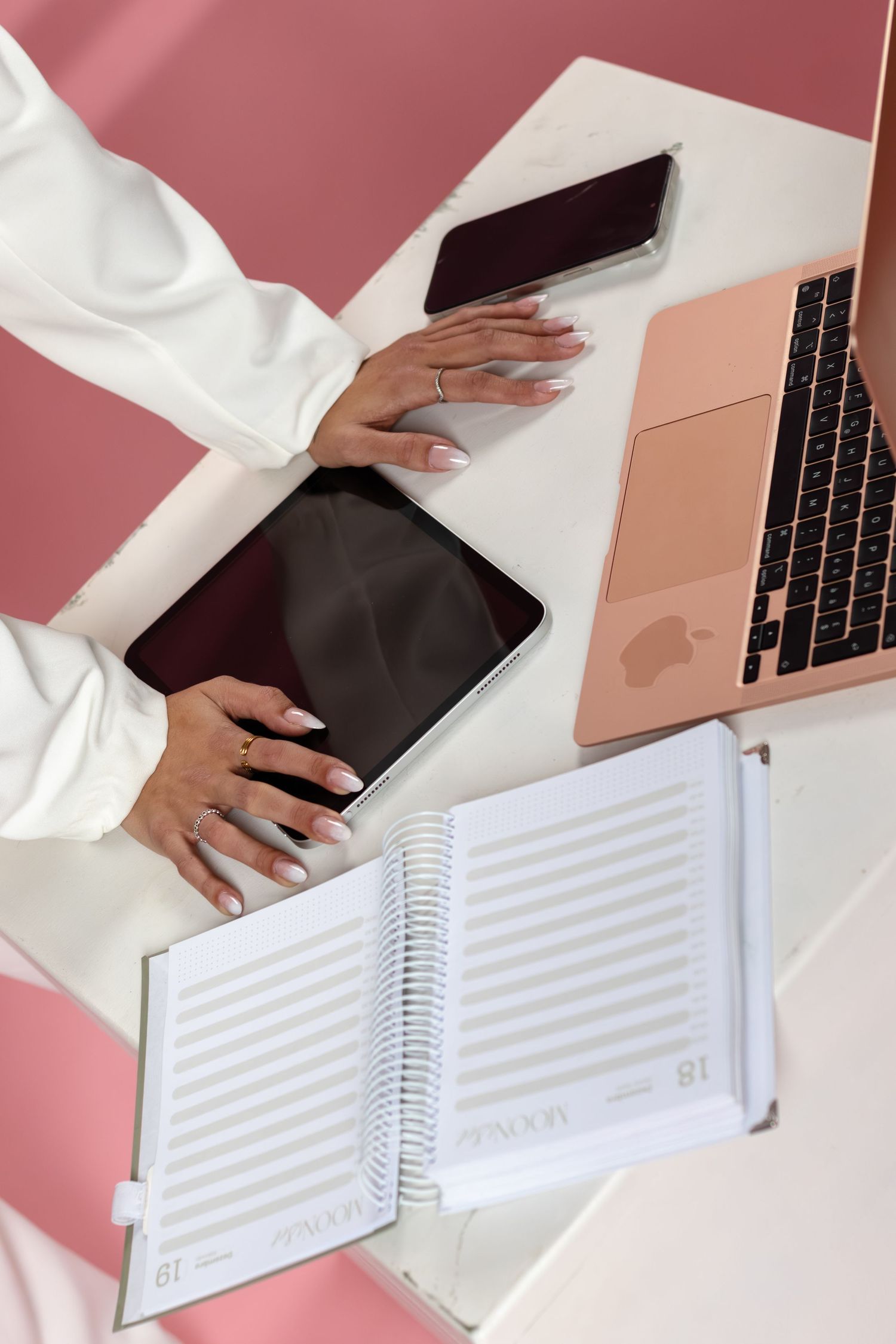 Hands in white sleeves working at desk with laptop, tablet, smartphone and spiral notebook on pink background.