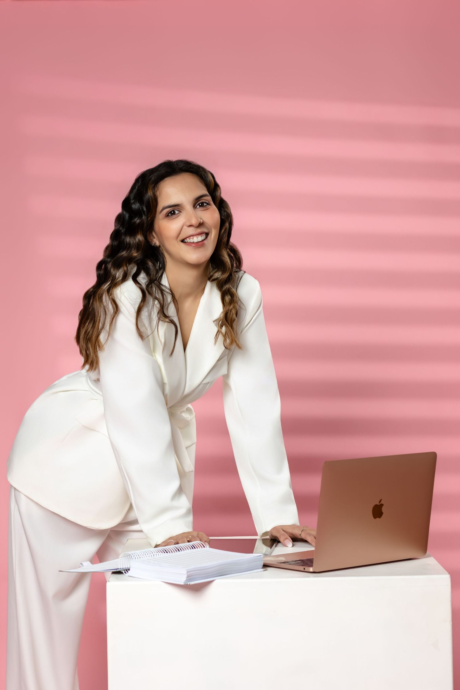 Professional in white business attire works on laptop against pink background with confident expression.