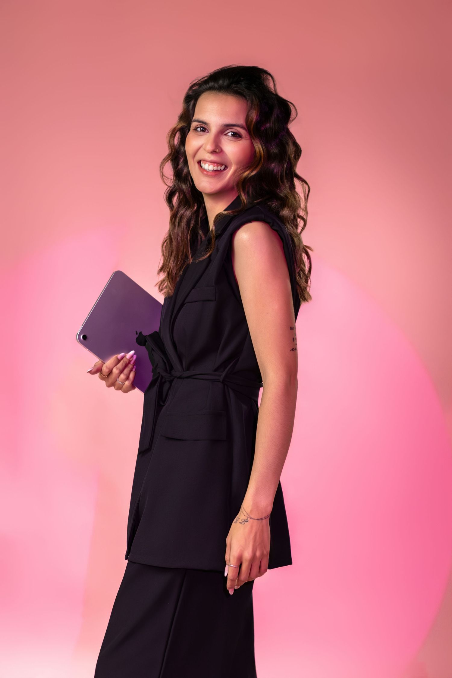 Professional portrait of someone in a sleek black outfit holding a tablet against a pink gradient background.