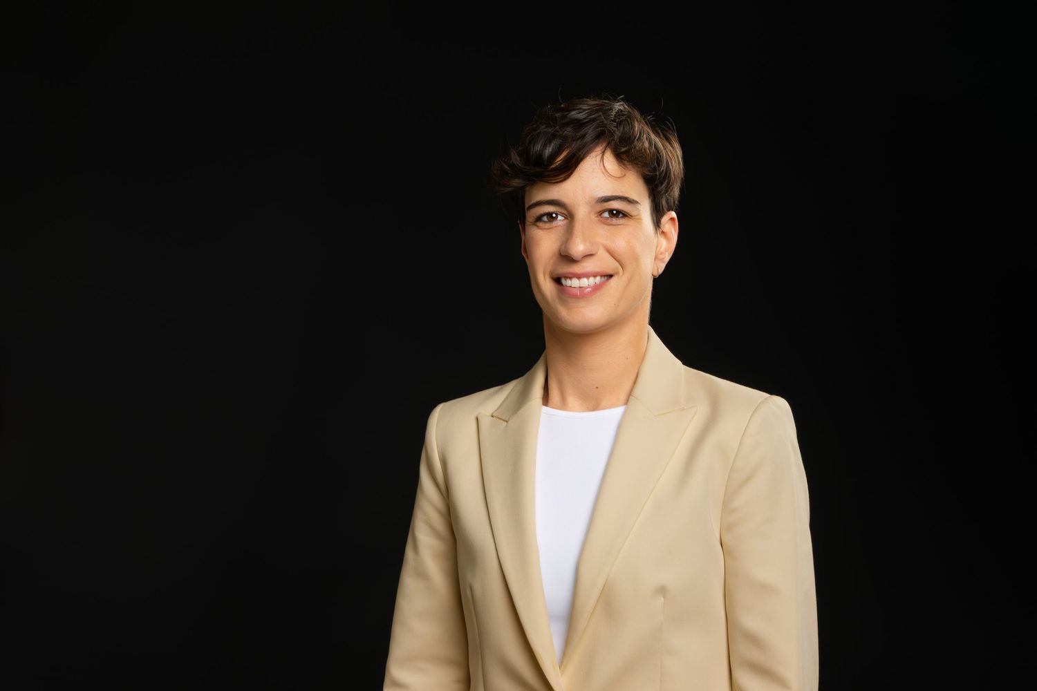 A person wearing a beige suit jacket and white shirt poses against a dark background.