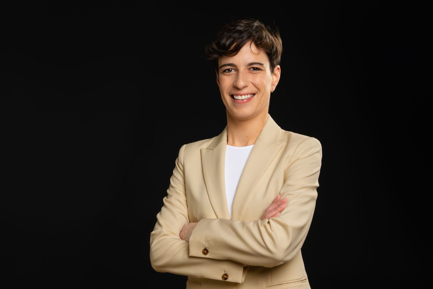 Professional headshot of someone wearing a beige blazer with arms crossed and a friendly smile against a dark background.