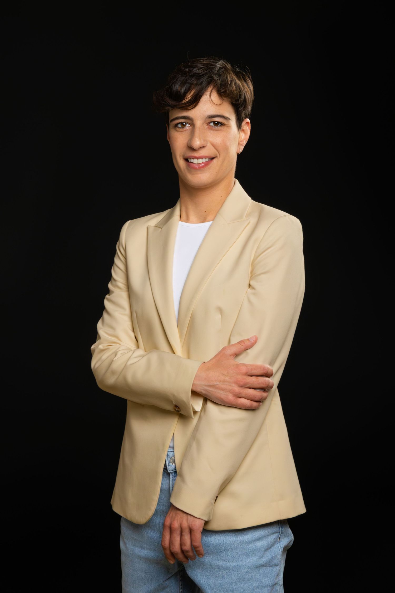 Professional portrait of someone wearing a beige blazer and jeans against a black background.