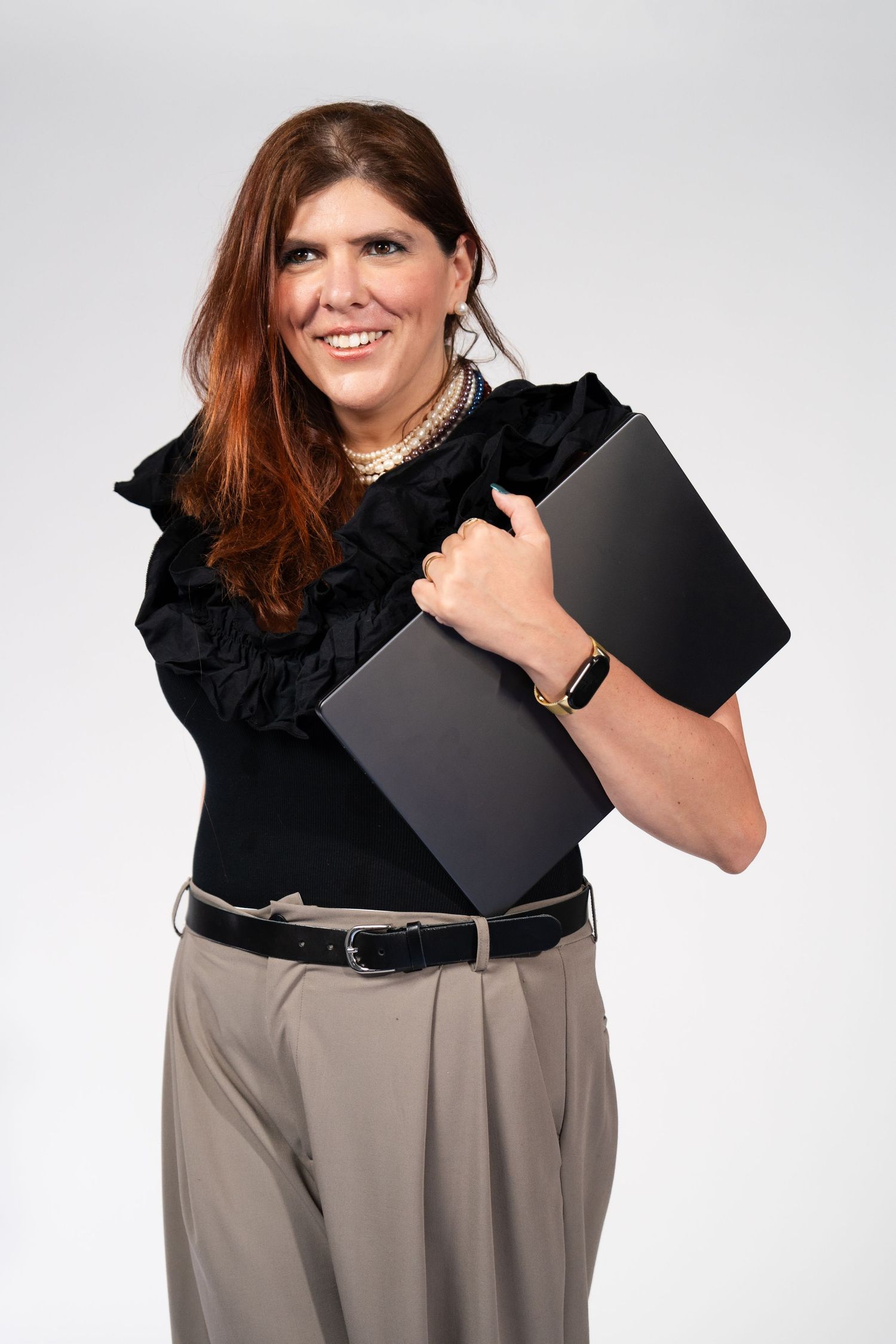 Woman in black top and gray pants holding black folder in professional studio poses.