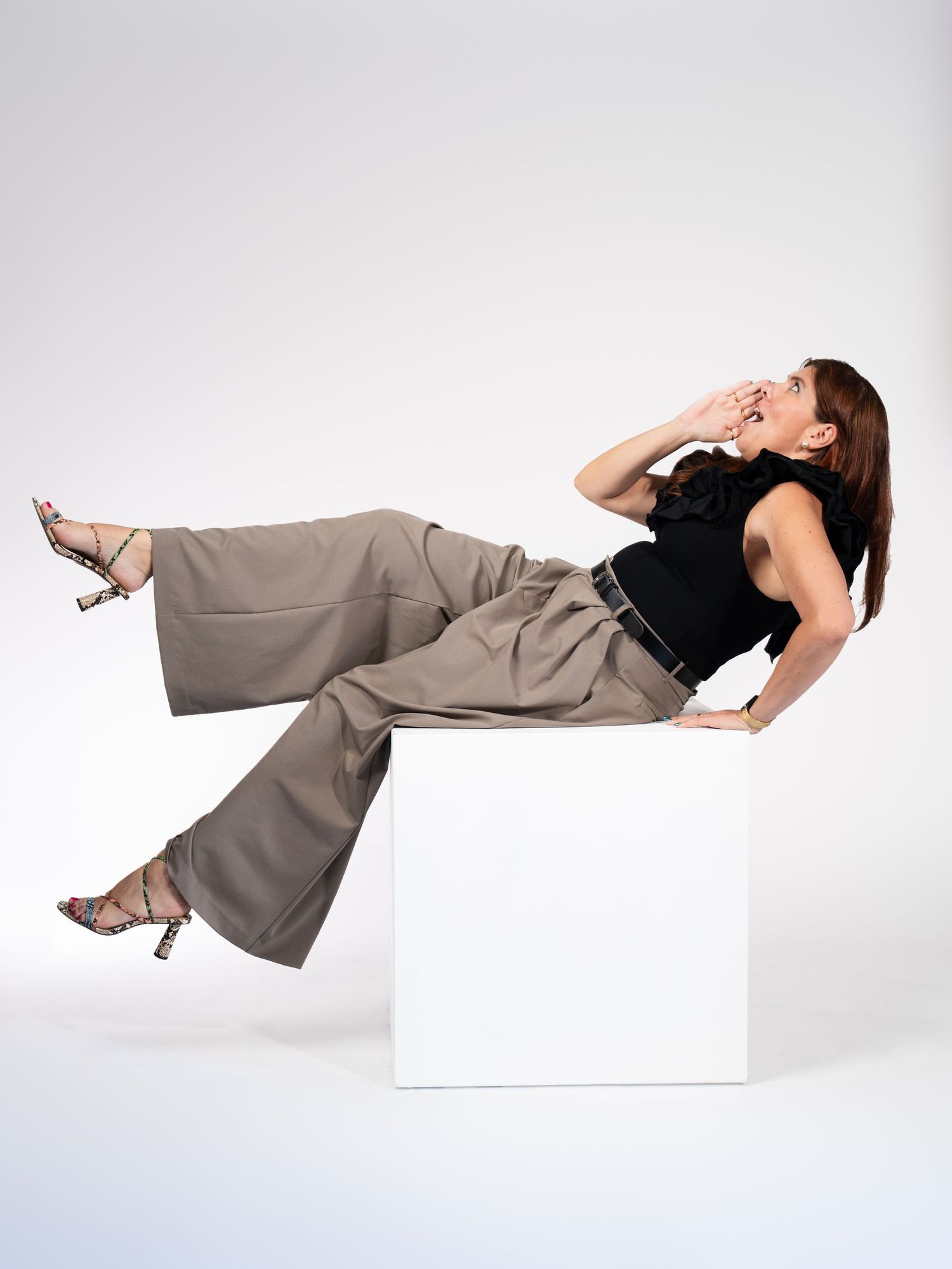 Model in black sleeveless top and gray pants posing on white platform in studio setting.