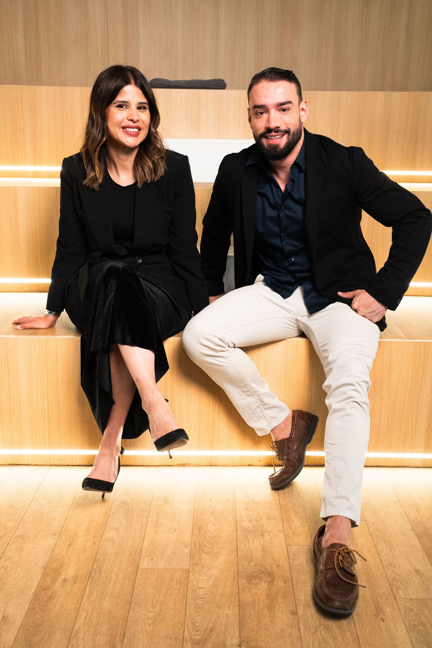 Two people in black attire posing playfully on wooden floor with warm lighting.