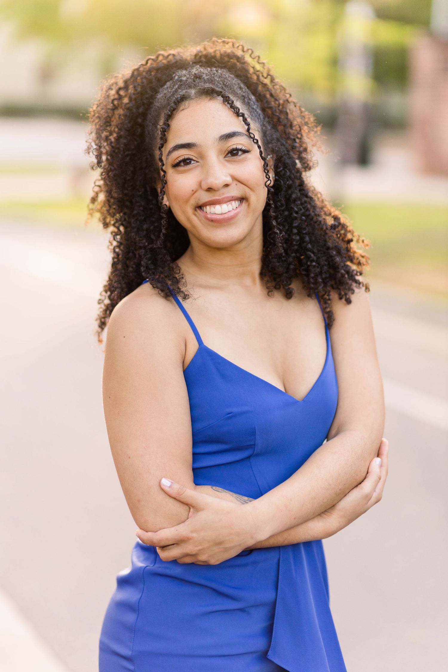 A person in a royal blue dress poses outdoors with natural curly hair and a bright smile.