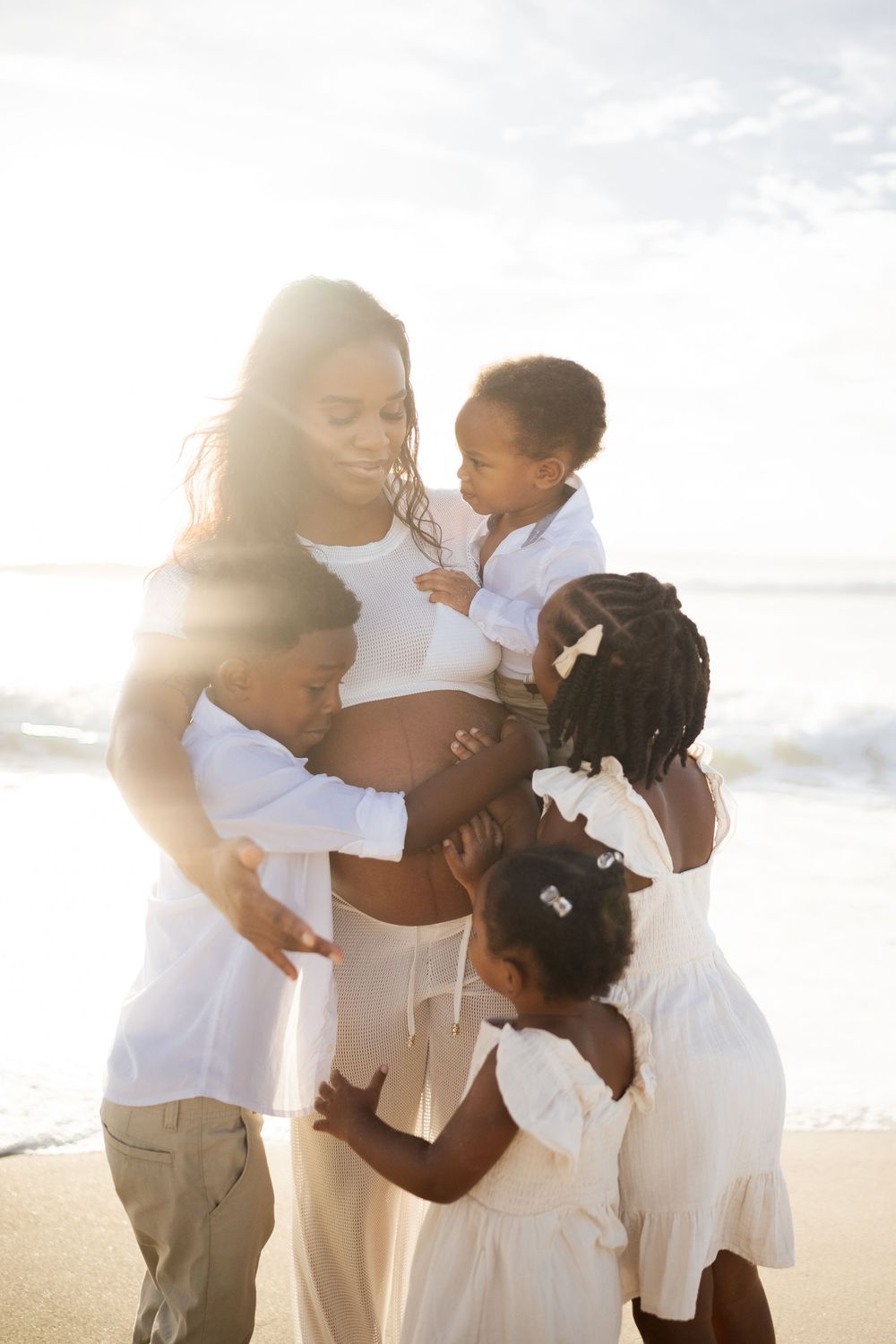 Pregnant Black mother embraced by her three young children on a sunny beach, all wearing white outfits.