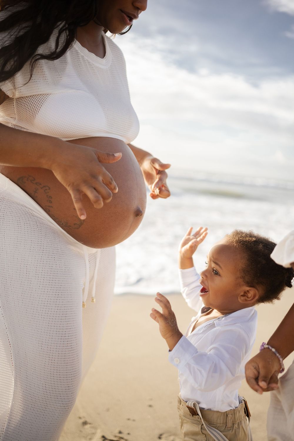 Pregnant woman in white dress on beach with young child reaching toward her baby bump on a sunny day.