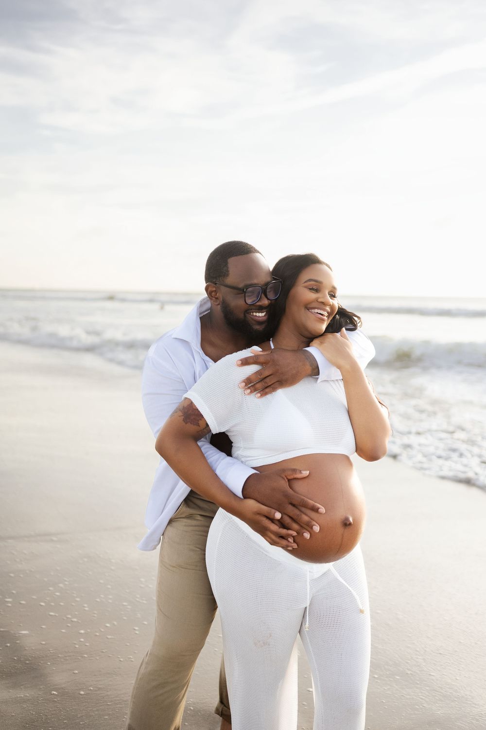 Happy couple on beach, pregnant woman in white outfit embraced by smiling man, ocean waves in background.
