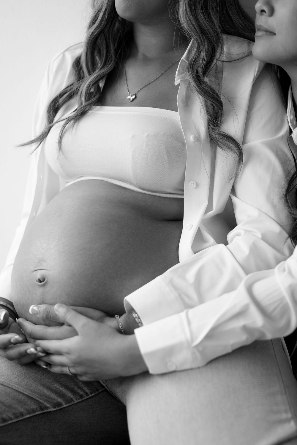 Black and white photo of a pregnant woman in white shirt and bra, with partner embracing her baby bump.