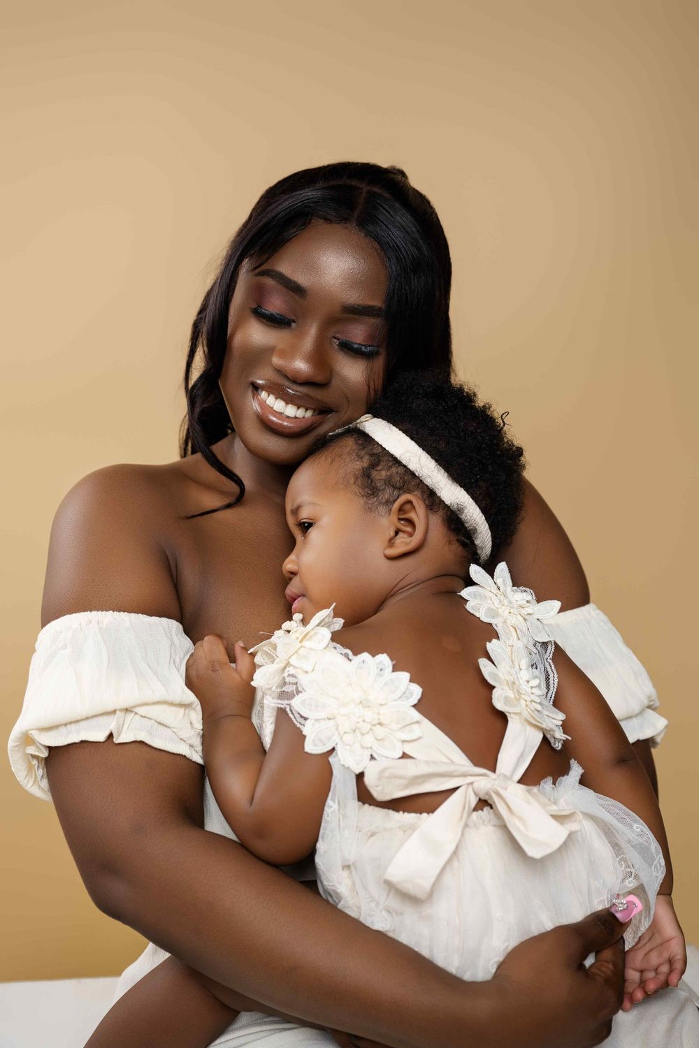 Mother and baby in matching white outfits share tender moment against beige background.