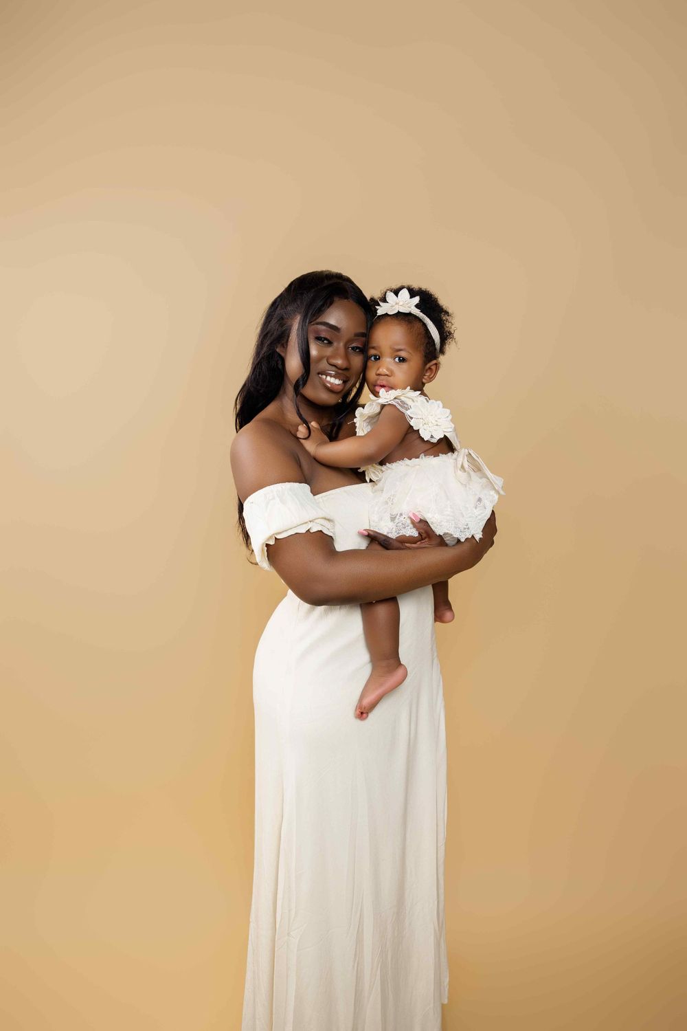 Mother and baby daughter in coordinating white dresses pose against warm neutral backdrop.