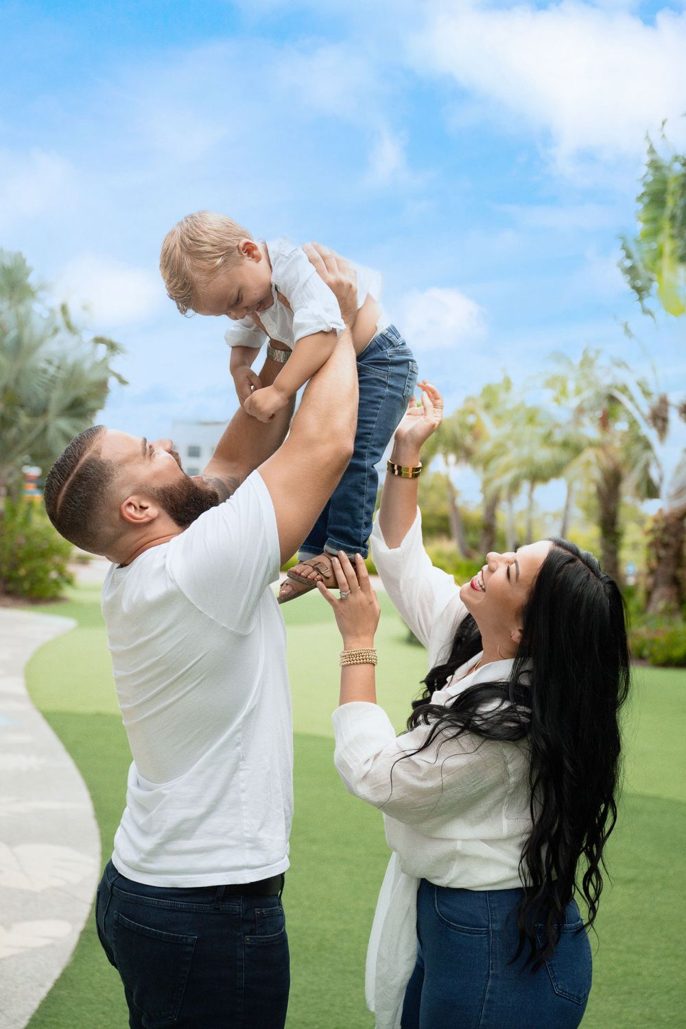 Family plays together outdoors as parents lift child in the air against blue sky and palm trees.