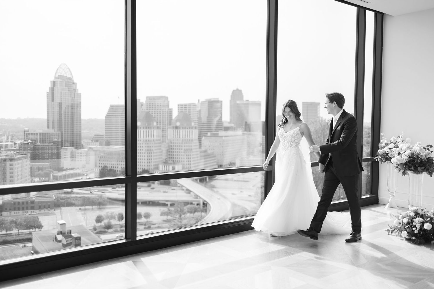 A black and white wedding photo with a couple standing by large windows overlooking a city skyline at The View Mt Adams.