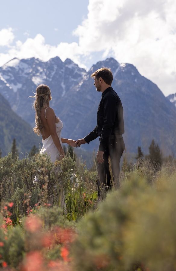 A couple in wedding attire holds hands in a wildflower meadow with snow-capped Grand Teton mountains behind them.