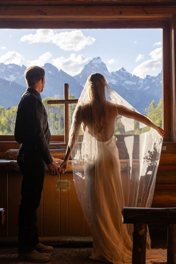 Bride and groom hold hands gazing at Grand Teton mountain views through chapel window on their wedding day.