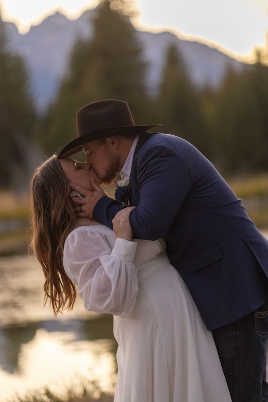 A romantic couple embraces and kisses at sunset by a serene lake with golden light reflecting on the water.