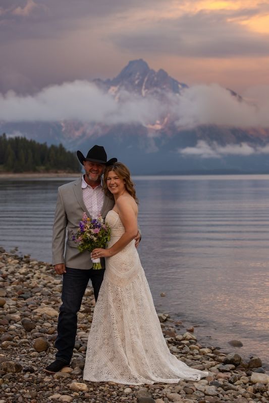 Couple in wedding attire poses by mountain lake at sunset, bride in lace gown holding bouquet, groom in cowboy hat.