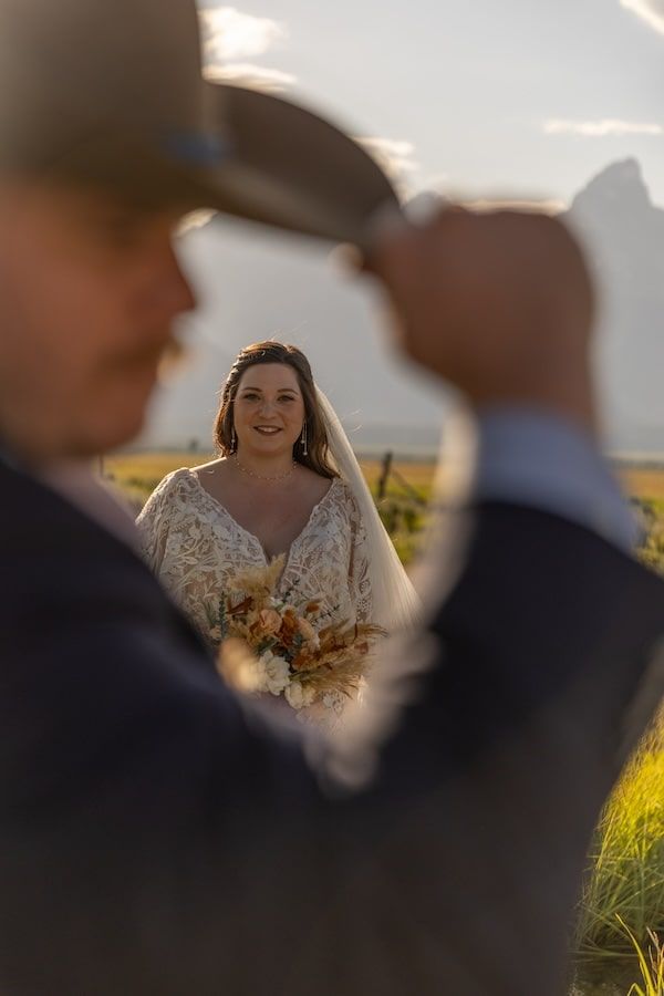 A bride in a lace wedding dress with bouquet viewed through silhouetted figures during an outdoor golden hour ceremony.