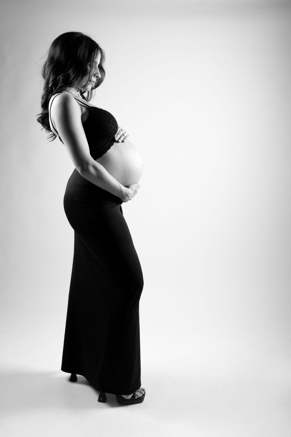 Black and white maternity portrait showing silhouette of pregnant woman in long dress against light background.