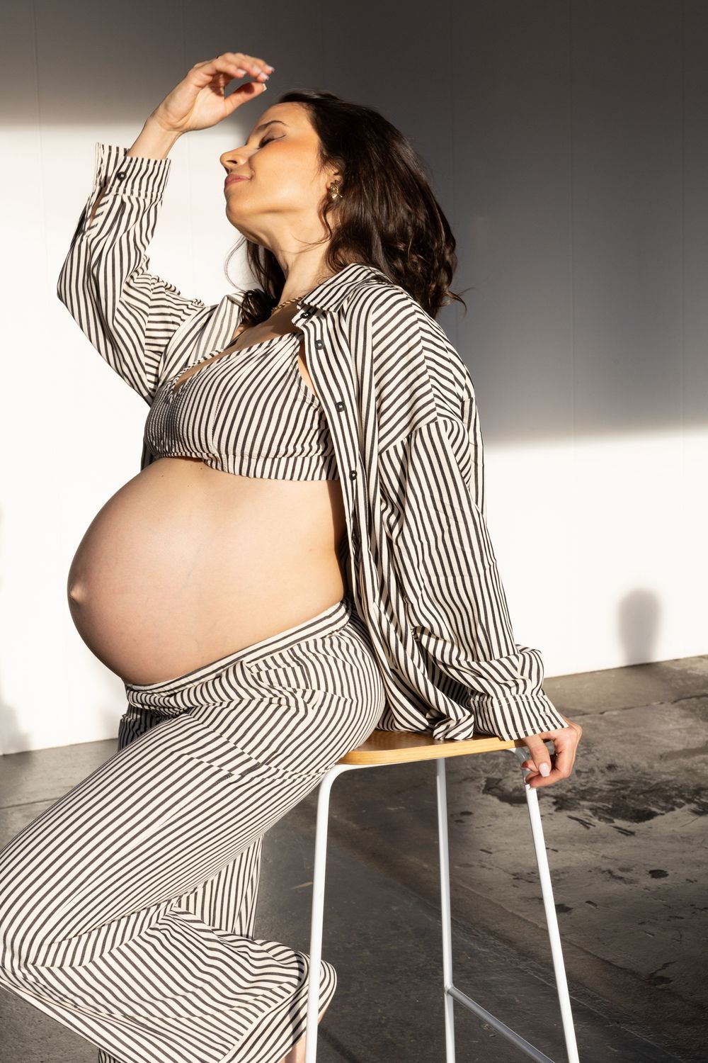 Pregnant person in striped loungewear sits on chair in dramatic lighting, showcasing baby bump in artistic pose.