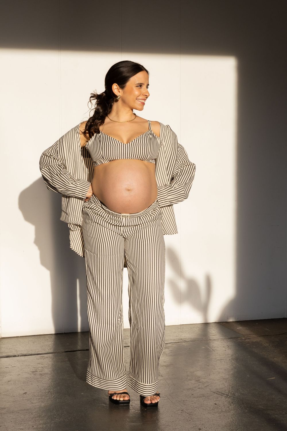 A pregnant woman in striped loungewear poses against a white wall with dramatic shadows creating artistic silhouettes.