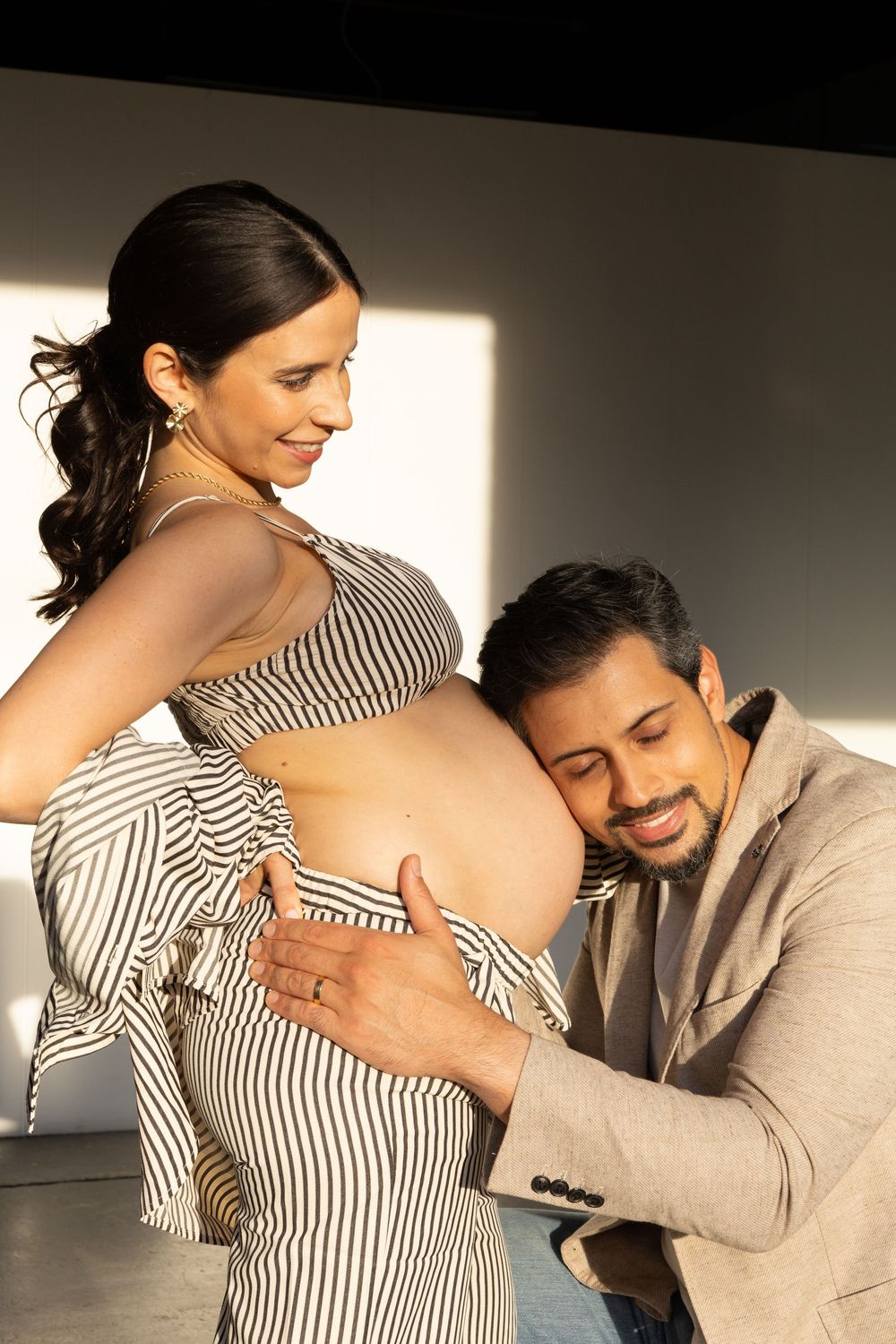 Expecting couple shares intimate moment as they pose for maternity photo in striped dress and beige blazer.