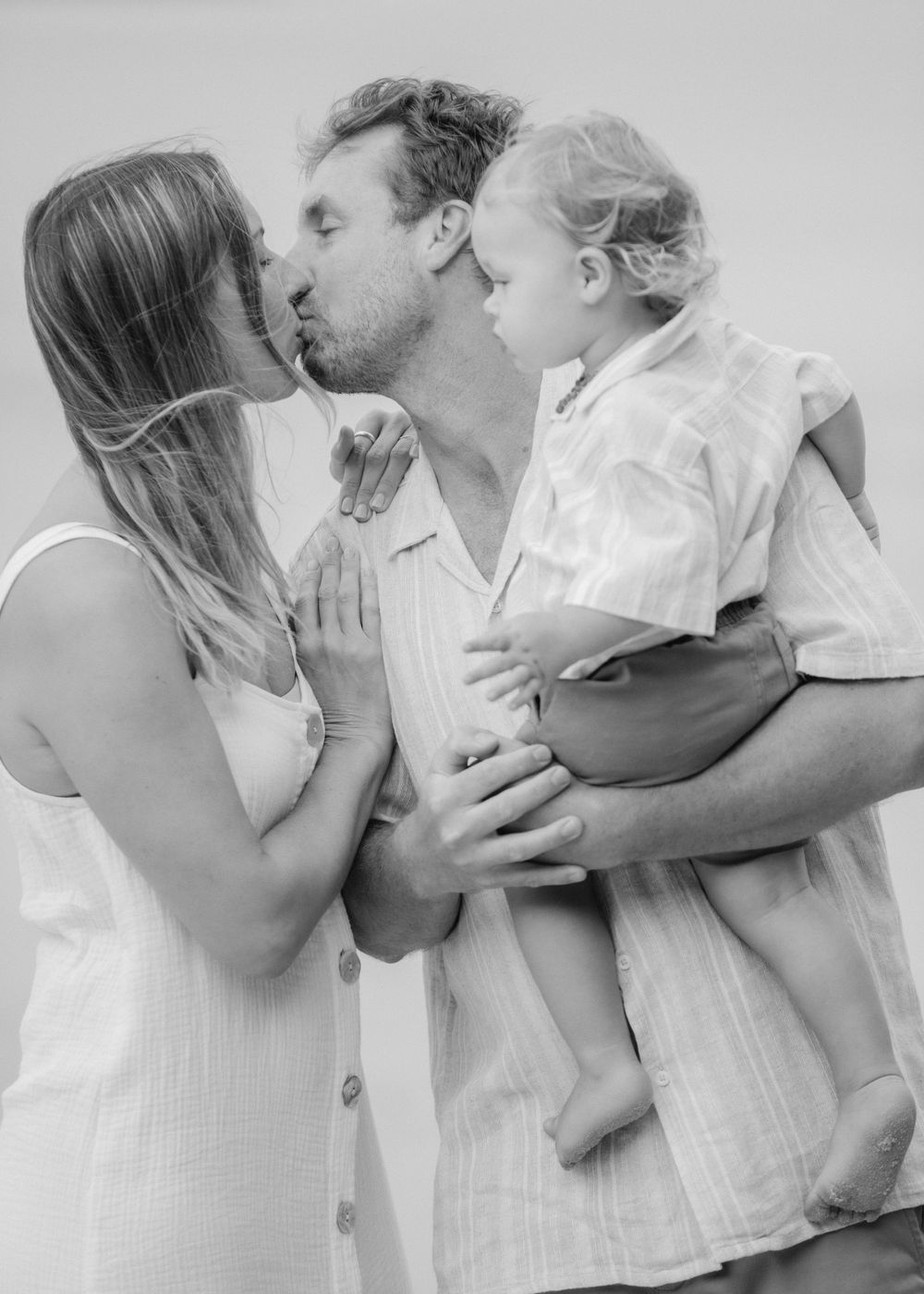 A tender black and white family portrait showing parents sharing a kiss while holding their young child.