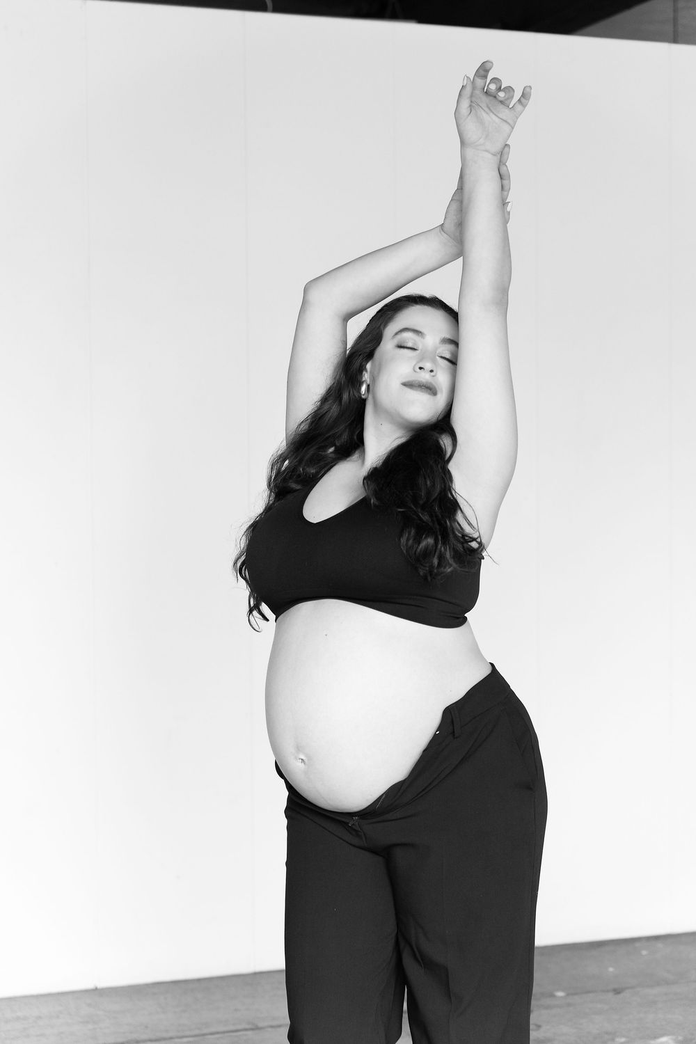 A series of black and white maternity photos showing a pregnant figure in dynamic dance-like poses against a white wall.