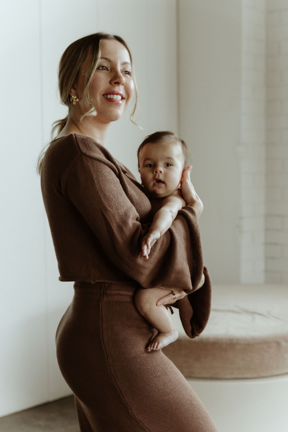 A parent wearing a brown dress holds a baby in matching brown tones while standing near a bright window.