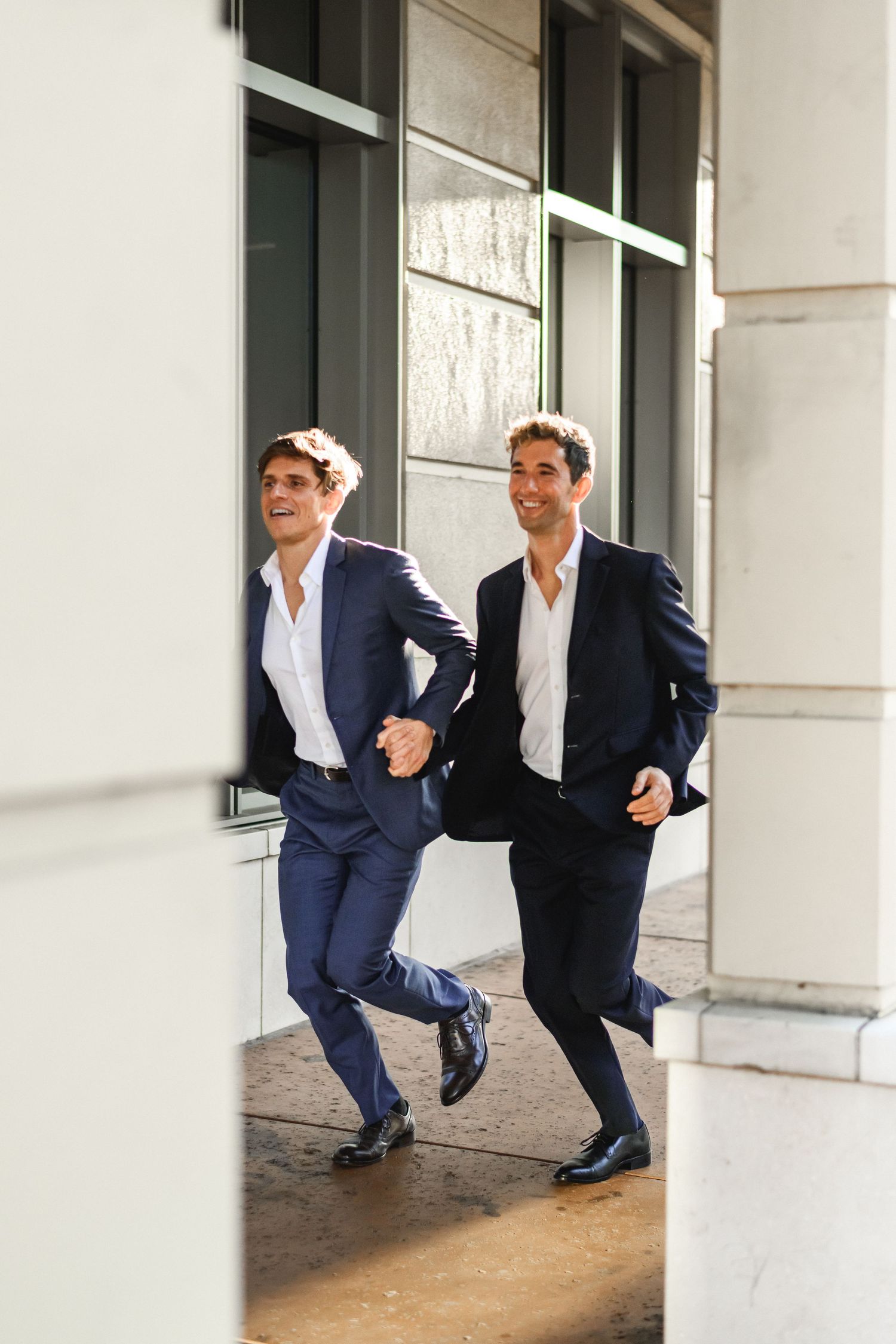 Two men in navy suits laughing and running together outside a modern building.