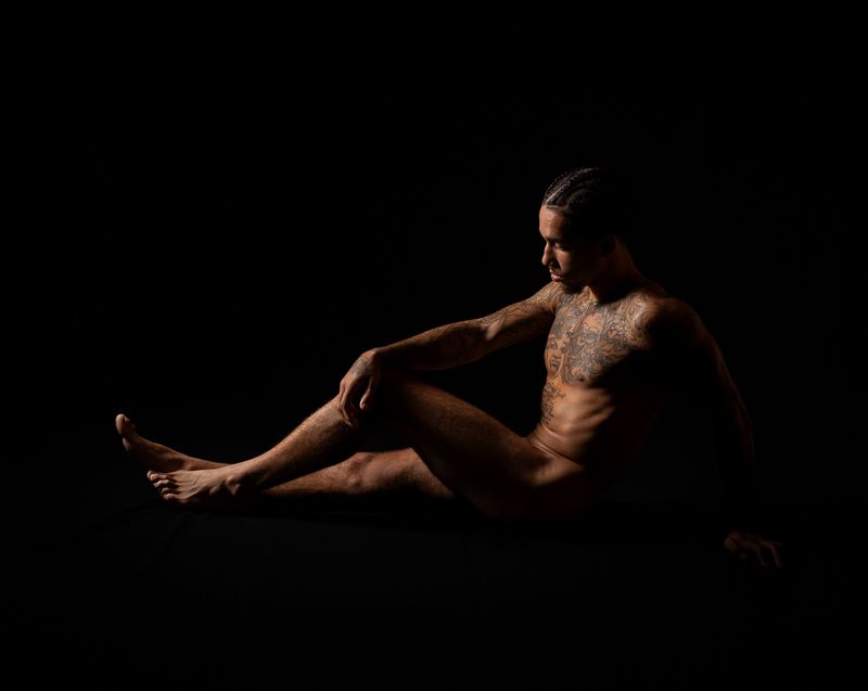 Dramatic low-key portrait of muscular figure seated in darkness with artistic shadows and lighting.