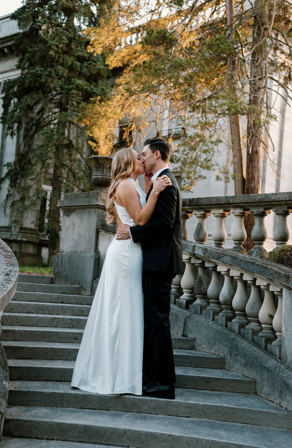 A couple shares a romantic kiss on stone steps surrounded by autumn trees and decorative white balustrades.