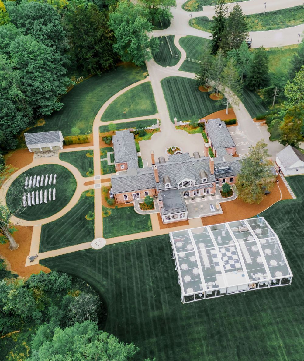 Aerial view of a white event tent and seating setup on a manicured lawn surrounded by trees and pathways.