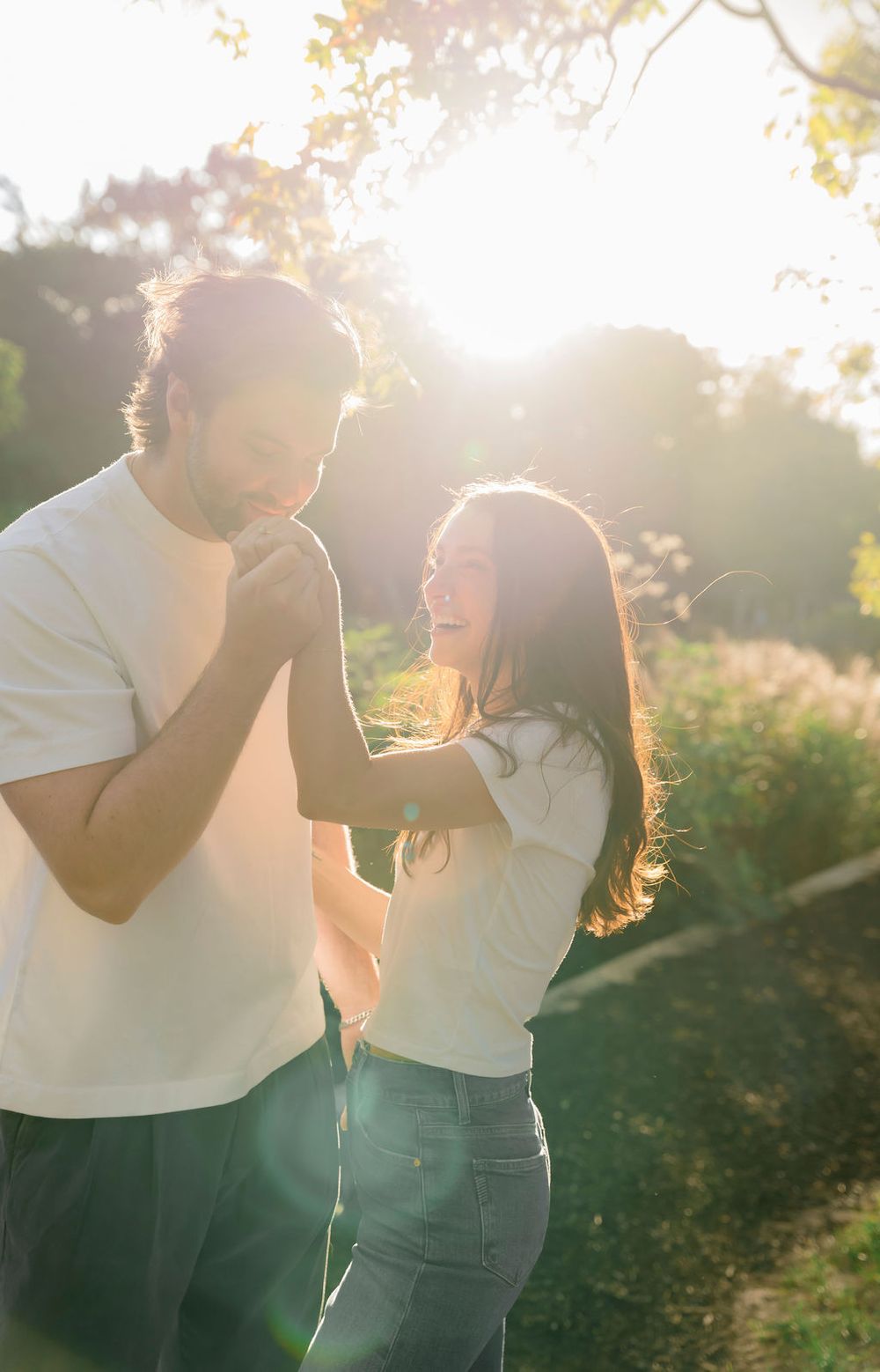 A couple shares a joyful moment outdoors during golden hour with sunlight streaming through trees in the background.