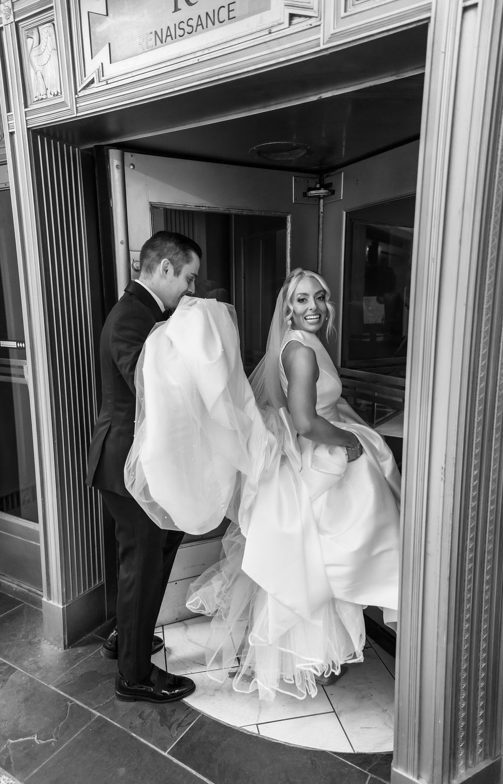 A bride in a white dress playfully enters through a doorway marked 'Naissance' with her companion in a black suit.